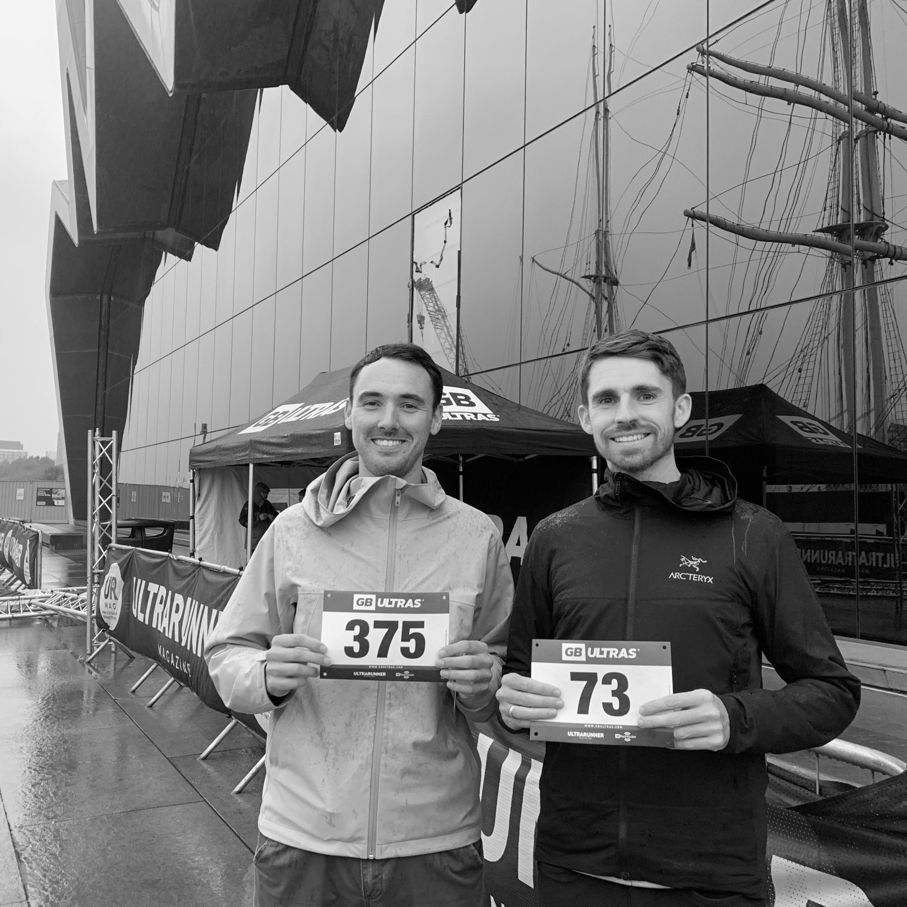 Two runners holding race numbers in front of a large ship.